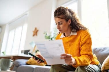 Households-woman seated in a cozy, well-lit living room reviewing a document, likely a bill, while holding a smartphone