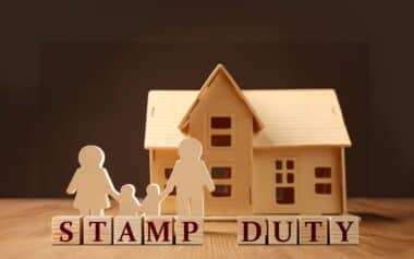 The image features a wooden house model with figures representing first-time buyers, and the words STAMP DUTY spelled out in wooden blocks.