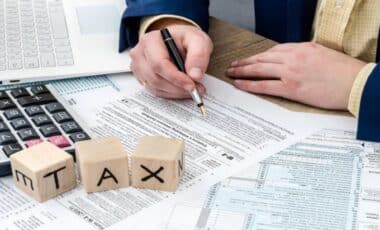 A Person Signing Personal Tax Allowance Documents at a desk with a calculator and Wooden Blocks Spelling "TAX"