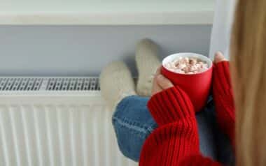 Energy Bills -A person dressed in a red sweater and jeans is sitting near a radiator with their feet