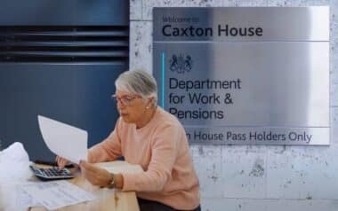 An older woman sits at a desk, reviewing documents with a DWP "Department for Work & Pensions" DWP sign in the background.