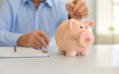 Senior Man Saving Money and Putting Coin in Cute Shiny Piggy Bank Standing on Desk