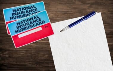National Insurance number cards placed on a dark wooden table next to a blank sheet of paper and a metallic blue pen