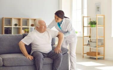Nurse Helping Senior Man to Stand up from Sofa in Modern Hospital or Geriatric Rehabilitation Center
