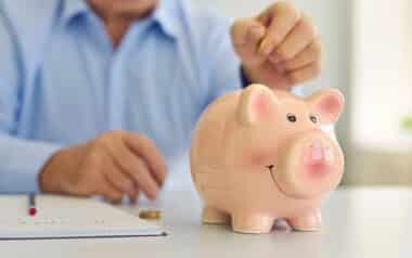 Senior Man Saving Money and Putting Coin in Cute Shiny Piggy Bank Standing on Desk