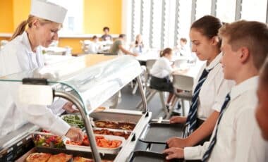 A lunch lady serves hot meals to students dressed in school uniforms along a cafeteria line.