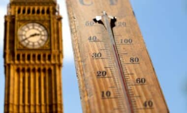 A wooden outdoor thermometer showing a high temperature of around 40°C (104°F) with Big Ben blurred in the background under a clear blue sky.