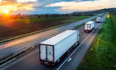 semi-trucks with large white trailers driving down a multi-lane highway at sunset
