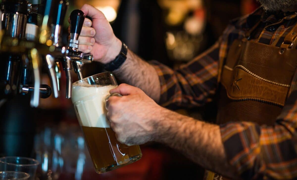 A bartender wearing an apron pours draft beer from a tap into a glass mug inside a brewery.