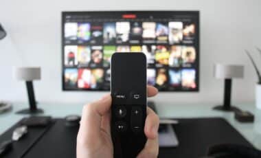 A person holding a black TV remote control in the foreground, with a flat-screen TV displaying a grid of media or movie thumbnails mounted on a wall in the background.