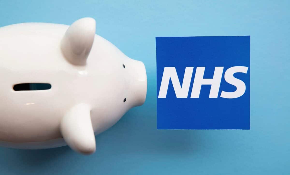 A white piggy bank sits on a light blue surface facing a blue square card featuring the white NHS logo.