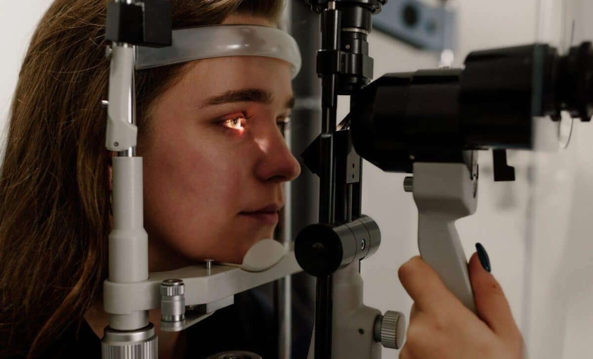 A woman has her head resting on a chin rest during an eye exam while a medical professional operates a slit lamp machine.