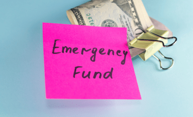 "Emergency Fund" written over a Pink Post-it Next to a Dollar Bill Clipped with a Yellow Paper Clip