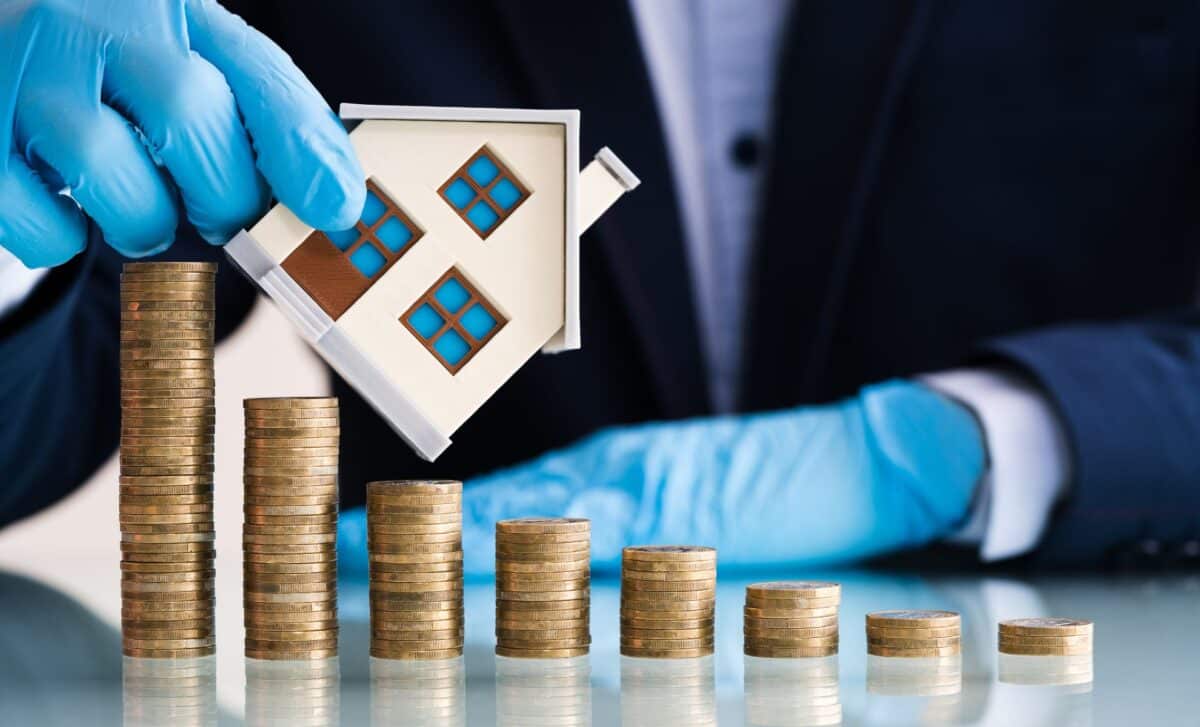 Gloved Hands Holding A Miniature House Over A Decreasing Set Of Coins To Symbolise UK House Prices Decline