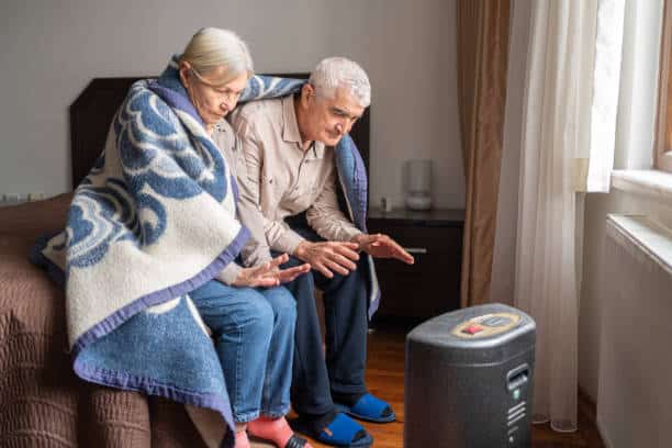 pensioners-Senior Couple Sitting on a Sofa with a Blanket Wrapped Around Them. They Appear to be Warming their Hands Near a Small Heater.