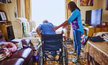 Carers- An Elderly Person, Seated In A Wheelchair, Is Being Assisted By A Carer