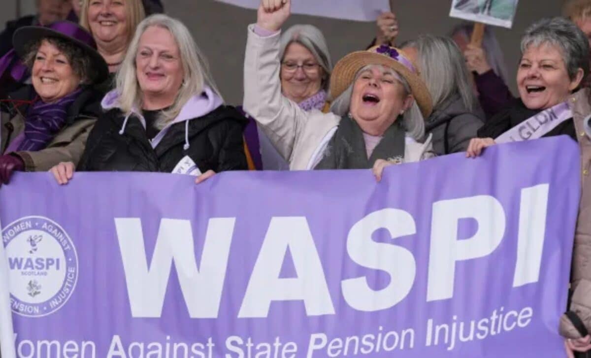 A Group Of Women Gathered Together Holding A Banner That Reads WASPI Women (Women Against State Pension Injustice).