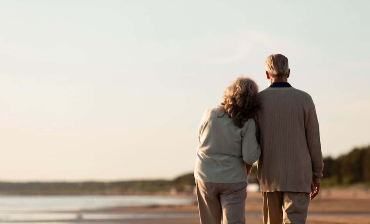 Two Retirees Standing Closely Facing A Serene Beach Under A Bright Sky