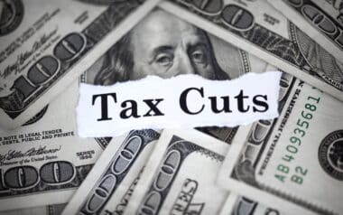 close-up view of U.S. $100 bills arranged in the background, with the words Tax Cuts prominently displayed on a torn piece of white paper layered over the money