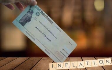 A hand holds a U.S. Treasury stimulus check labeled Relief Program, with Scrabble tiles spelling INFLATION in the foreground.