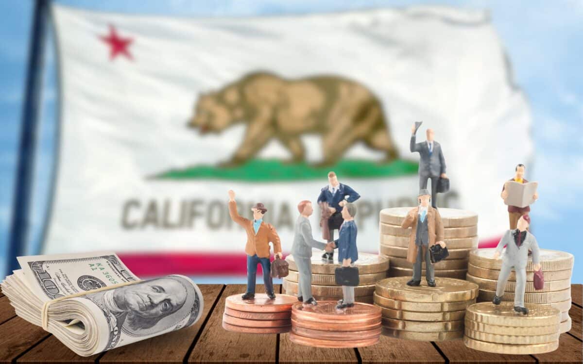 miniature business figures standing on stacks of coins and US dollar bills, with the California state flag blurred in the background