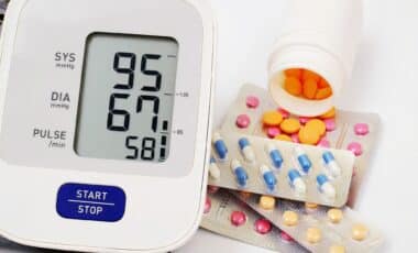 A digital blood pressure monitor displaying a reading of 9567 sits alongside various colorful pills and an open medicine bottle on a white surface.